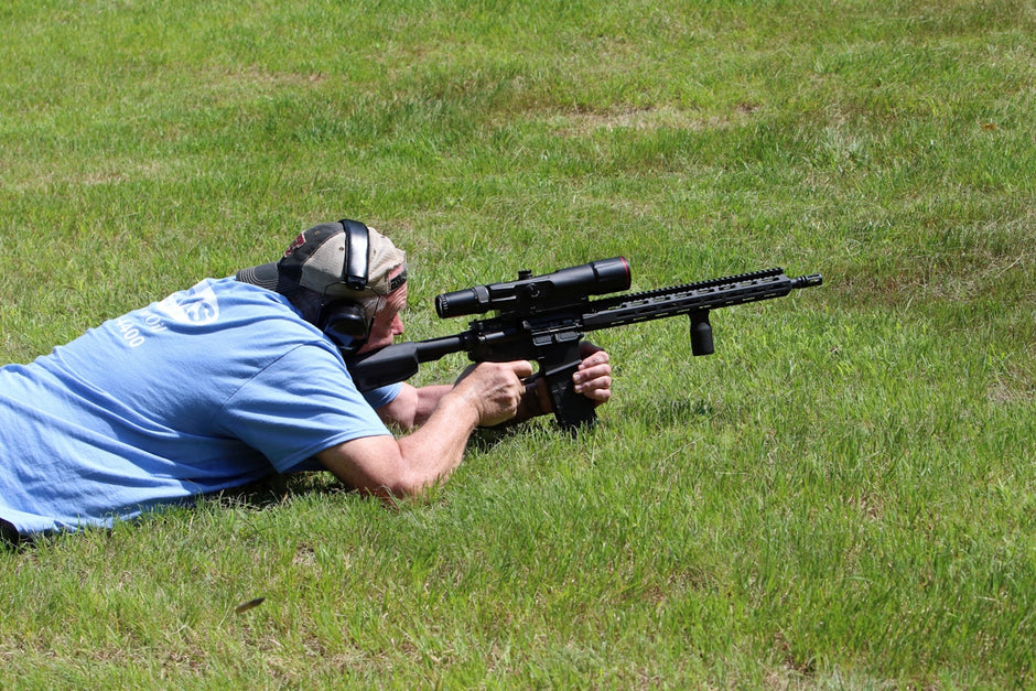 Man lies prone, aiming a rifle on grass.