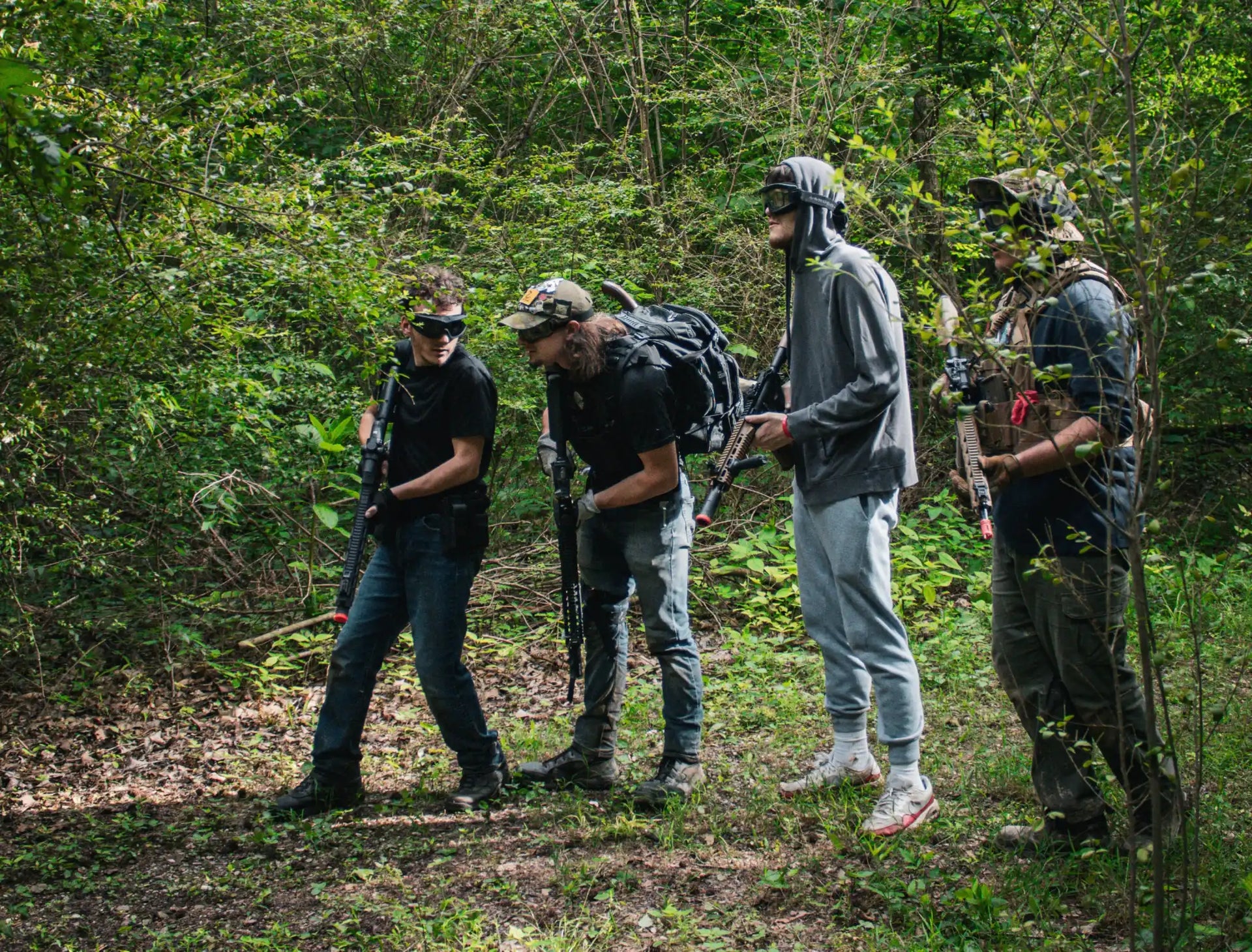 Four people in a wooded area, equipped with protective eyewear and what appear to be airsoft guns and tactical gear.